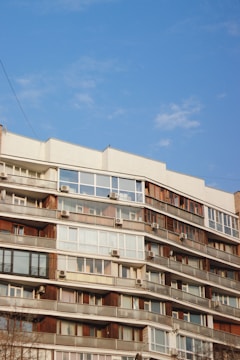 A multi-story residential building features numerous balconies with windows and air conditioning units. The structure is characterized by a mix of wood and glass elements, and sits against a clear blue sky.