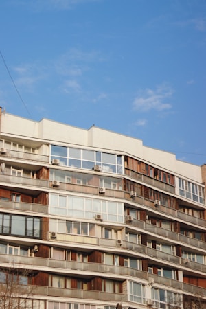 A multi-story residential building features numerous balconies with windows and air conditioning units. The structure is characterized by a mix of wood and glass elements, and sits against a clear blue sky.