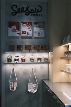 A display features coffee products and accessories on a wall with a pegboard background. Shelves hold multiple coffee packets and bags with a minimalist design. Two fabric shoulder bags hang from the pegboard.