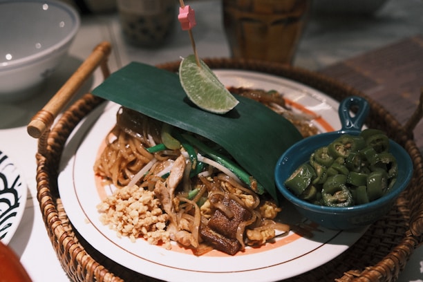 A plate of pad thai garnished with a slice of lime on a leaf, accompanied by chopped green chilies in a small blue bowl. The dish includes noodles, peanuts, and pieces of chicken or tofu, placed on a round woven basket tray.