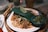 A plate of pad thai garnished with a slice of lime on a leaf, accompanied by chopped green chilies in a small blue bowl. The dish includes noodles, peanuts, and pieces of chicken or tofu, placed on a round woven basket tray.