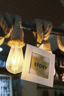 A glowing light bulb hangs next to a piece of rope wrapped around a metal bar. A square card with the word 'STORY' printed on it hangs beside the bulb, partially illuminated by its light. The background appears to be the interior of a room with blurred details, suggesting a cozy atmosphere.