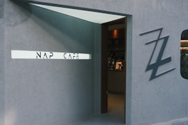 An entrance to a modern cafe with a minimalistic design featuring a sign that reads 'NAP CAFE'. The entrance is made of a muted gray concrete structure with a sleek, wooden door partially open. In the interior, some furniture and decor can be faintly seen. There is a stylized 'ZZ' logo mounted on the gray wall next to the entrance.