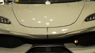 Close-up of the front of a sleek, modern car with a glossy finish. The design features smooth curves and a prominent logo at the center of the hood. The headlights have an angular, futuristic shape, and the grille below adds a sporty touch.