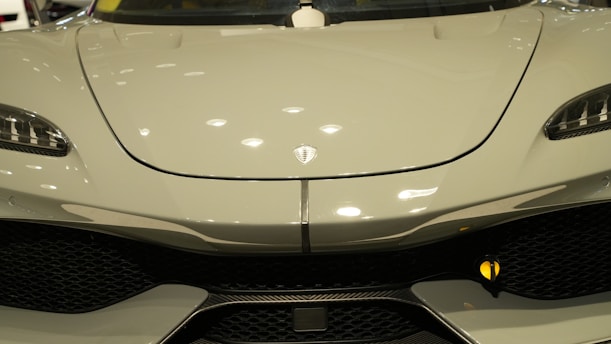 Close-up of the front of a sleek, modern car with a glossy finish. The design features smooth curves and a prominent logo at the center of the hood. The headlights have an angular, futuristic shape, and the grille below adds a sporty touch.