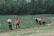 A group of six people are working together on a patch of land, engaging in agricultural activities. They are dressed in various colors with some donning traditional hats. The background consists of dense green foliage and trees, with a marshy area or river behind the workers. The scene suggests a rural setting with natural surroundings.