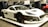 A sleek, modern electric hypercar with a white body and black accents displayed at a car show. The car features aerodynamic styling, carbon fiber elements, and large black rims with red detailing. Bright lights reflect off its smooth surface, highlighting its futuristic design.