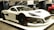 A sleek, modern electric hypercar with a white body and black accents displayed at a car show. The car features aerodynamic styling, carbon fiber elements, and large black rims with red detailing. Bright lights reflect off its smooth surface, highlighting its futuristic design.