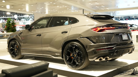 A sleek, modern SUV is showcased indoors, likely at an auto show. It features a glossy metallic gray finish and distinctive black rims. The rear lights are sharply designed, and the vehicle is positioned on a raised platform with soft, ambient lighting.