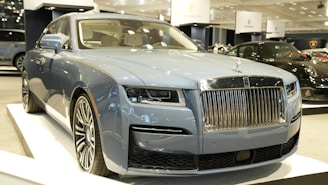A luxury vehicle in a showroom setting, with a prominent front grille and sleek design. The environment is well-lit, showcasing multiple high-end cars on display.