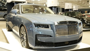 A luxury vehicle in a showroom setting, with a prominent front grille and sleek design. The environment is well-lit, showcasing multiple high-end cars on display.