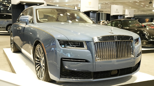 A luxury vehicle in a showroom setting, with a prominent front grille and sleek design. The environment is well-lit, showcasing multiple high-end cars on display.