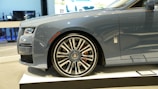 A luxury car is showcased indoors on a platform. The vehicle has a sleek, metallic gray exterior with a prominent, stylish wheel design featuring a distinctive emblem. The setting appears to be a showroom with a modern interior.