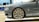 A luxury car is showcased indoors on a platform. The vehicle has a sleek, metallic gray exterior with a prominent, stylish wheel design featuring a distinctive emblem. The setting appears to be a showroom with a modern interior.