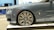 A luxury car is showcased indoors on a platform. The vehicle has a sleek, metallic gray exterior with a prominent, stylish wheel design featuring a distinctive emblem. The setting appears to be a showroom with a modern interior.