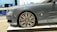 A luxury car is showcased indoors on a platform. The vehicle has a sleek, metallic gray exterior with a prominent, stylish wheel design featuring a distinctive emblem. The setting appears to be a showroom with a modern interior.