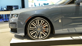 A luxury car is showcased indoors on a platform. The vehicle has a sleek, metallic gray exterior with a prominent, stylish wheel design featuring a distinctive emblem. The setting appears to be a showroom with a modern interior.