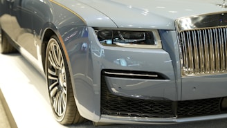 A close-up view of the front section of a luxury car featuring a sleek, metallic blue exterior. The image prominently displays the detailed headlight design, the intricate grille with a chrome finish, and a large, stylized wheel. The surface reflects ambient light, highlighting the car's smooth curves and high-end finish.