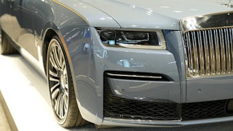 A close-up view of the front section of a luxury car featuring a sleek, metallic blue exterior. The image prominently displays the detailed headlight design, the intricate grille with a chrome finish, and a large, stylized wheel. The surface reflects ambient light, highlighting the car's smooth curves and high-end finish.