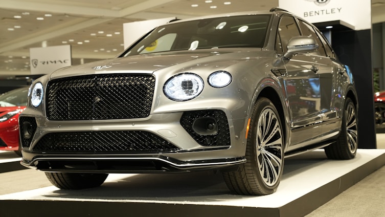 A luxury SUV car with a sleek, metallic silver finish is displayed on a platform in an indoor showroom. The car features distinctive, illuminated round headlights and a prominent grille. There are signs visible in the background, and other cars can be seen nearby.