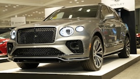 A luxury SUV car with a sleek, metallic silver finish is displayed on a platform in an indoor showroom. The car features distinctive, illuminated round headlights and a prominent grille. There are signs visible in the background, and other cars can be seen nearby.
