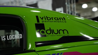 A close-up view of a vibrant green car with decals featuring the words 'Vibrant Performance' and 'Radium Engineering'. The car door window displays the text 'IS LOCKED'. The background suggests an indoor setting, possibly at a car show or exhibition.