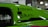 A close-up view of a vibrant green car with decals featuring the words 'Vibrant Performance' and 'Radium Engineering'. The car door window displays the text 'IS LOCKED'. The background suggests an indoor setting, possibly at a car show or exhibition.