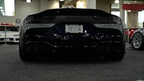 A sleek black Maserati sports car is showcased at an indoor car event with a custom license plate '7DESIGN'. The setting includes other cars in the background, visible through a brightly lit, spacious exhibition hall.