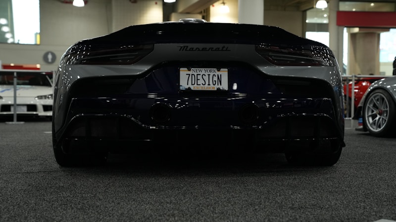 A sleek black Maserati sports car is showcased at an indoor car event with a custom license plate '7DESIGN'. The setting includes other cars in the background, visible through a brightly lit, spacious exhibition hall.
