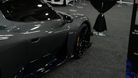 A sleek, dark-colored sports car is displayed indoors on a carpeted floor. The focus is on the side and front wheel of the vehicle, which features a glossy finish. Part of another vehicle is partially visible in the background, along with stanchions and a curtain divider.