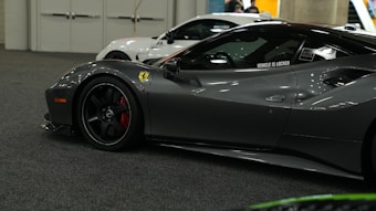 A sleek, metallic gray sports car is prominently displayed indoors on a carpeted floor. The car features a distinctive badge on the side, polished black rims, and a slightly open driver-side window with a decal that reads 'VEHICLE IS LOCKED'. In the background, parts of another white vehicle are visible, along with people and structural elements of the building.