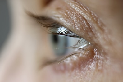 A close-up view of a human eye, focusing on the eyelashes and the texture of the skin around the eye. The image captures fine details such as the curvature of the eyelashes and the delicate creases and pores of the skin.
