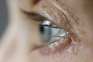 A close-up view of a human eye, focusing on the eyelashes and the texture of the skin around the eye. The image captures fine details such as the curvature of the eyelashes and the delicate creases and pores of the skin.