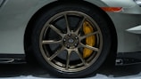 A close-up view of a car's wheel with bronze-colored alloy rims and a yellow brake caliper. The tire tread is visible with the brand name Dunlop inscribed on it. The car body is painted in a grayish-silver color, and part of the front bumper and a side marker light are also visible.