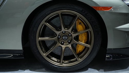 A close-up view of a car's wheel with bronze-colored alloy rims and a yellow brake caliper. The tire tread is visible with the brand name Dunlop inscribed on it. The car body is painted in a grayish-silver color, and part of the front bumper and a side marker light are also visible.