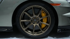 A close-up view of a car's wheel with bronze-colored alloy rims and a yellow brake caliper. The tire tread is visible with the brand name Dunlop inscribed on it. The car body is painted in a grayish-silver color, and part of the front bumper and a side marker light are also visible.