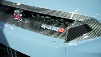 An image features the rear section of a sports car with carbon fiber detailing and a prominent Nismo branding. The emblem is red and silver, set against a backdrop of sleek, aerodynamic design elements typical of high-performance vehicles.