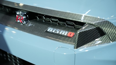 An image features the rear section of a sports car with carbon fiber detailing and a prominent Nismo branding. The emblem is red and silver, set against a backdrop of sleek, aerodynamic design elements typical of high-performance vehicles.