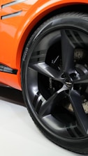 A close-up view of a car wheel featuring a sleek, metallic-gray rim with the brand name 'Pirelli' visible on the tire. The car's body is a vibrant orange with black accents, creating a sporty and modern aesthetic.