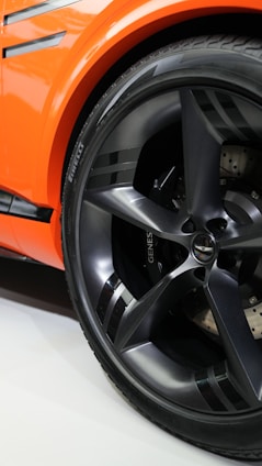 A close-up view of a car wheel featuring a sleek, metallic-gray rim with the brand name 'Pirelli' visible on the tire. The car's body is a vibrant orange with black accents, creating a sporty and modern aesthetic.