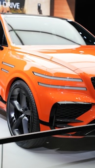 A vibrant orange sports car with sleek lines and aerodynamic features is displayed. The car has a modern design with distinctive black grille accents and large alloy wheels. It is positioned on a white showroom floor with soft lighting reflecting off its glossy surface.