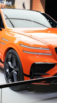 A vibrant orange sports car with sleek lines and aerodynamic features is displayed. The car has a modern design with distinctive black grille accents and large alloy wheels. It is positioned on a white showroom floor with soft lighting reflecting off its glossy surface.