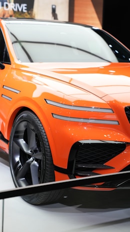 A vibrant orange sports car with sleek lines and aerodynamic features is displayed. The car has a modern design with distinctive black grille accents and large alloy wheels. It is positioned on a white showroom floor with soft lighting reflecting off its glossy surface.