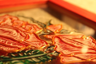 Close-up view of an embossed glass art piece featuring detailed red rose petals with golden outlines. The composition includes green leaves, adding contrast against the warm tones of the petals. The artwork is framed in a rectangular shape.