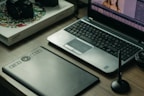 A workspace featuring a laptop with photo-editing software open, a graphics tablet, a digital pen in a holder, and a box labeled 'Wacom Intuos Pro.' Additionally, there's a DSLR camera resting on the box.