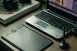 A workspace featuring a laptop with photo-editing software open, a graphics tablet, a digital pen in a holder, and a box labeled 'Wacom Intuos Pro.' Additionally, there's a DSLR camera resting on the box.