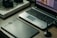 A workspace featuring a laptop with photo-editing software open, a graphics tablet, a digital pen in a holder, and a box labeled 'Wacom Intuos Pro.' Additionally, there's a DSLR camera resting on the box.
