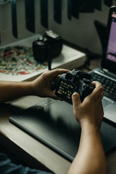 A person is holding a DSLR camera in their hands, examining the settings displayed on the screen. A digital drawing tablet is placed in front of them on the desk, next to a laptop with a visible screen showing editing software. In the background, there's a camera lens on the table, and a colorful pattern is partially visible.