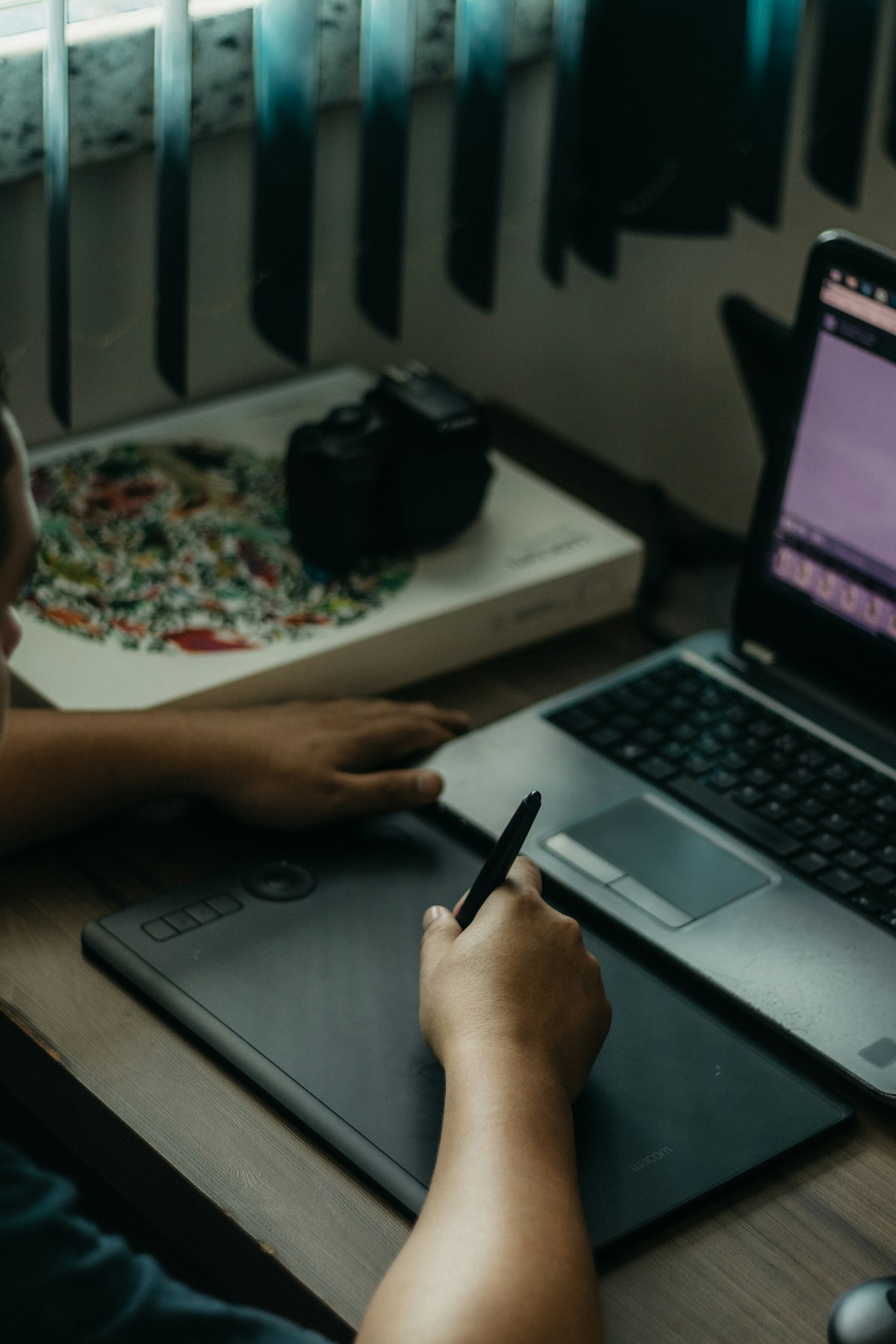 Personne dessinant au stylet sur une tablette graphique noire posée sur un bureau en bois, à côté d'un ordinateur portable ouvert