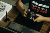A person is seated at a desk, holding a DSLR camera. On the desk, there is a laptop, a graphic tablet, and a stylus pen. The person wears a dark shirt with the word 'RESILIÊNCIA' printed on it. There is also a piece of artwork on the desk featuring floral designs.